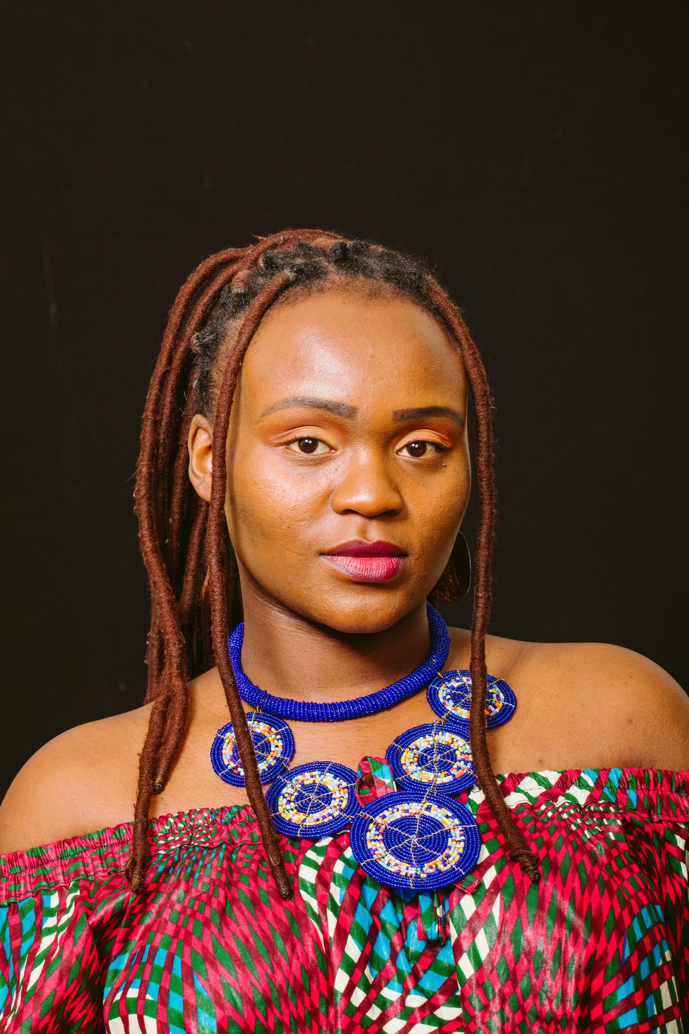 Woman braids + blue statement necklace, dark bg, polished cultural portrait.